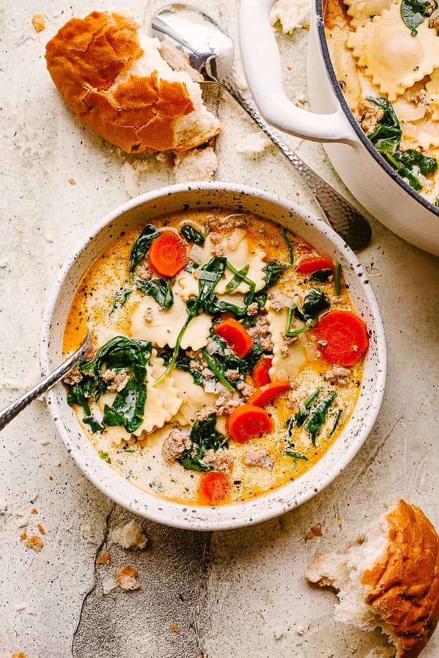 A bowl with creamy ravioli soup. Torn up bread is placed near the bowl.