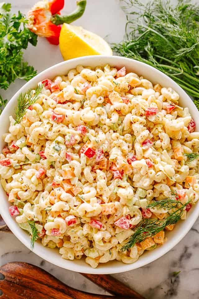 Homemade macaroni salad in a large bowl, with herbs, lemon halves, and serving spoons arranged on the work surface.