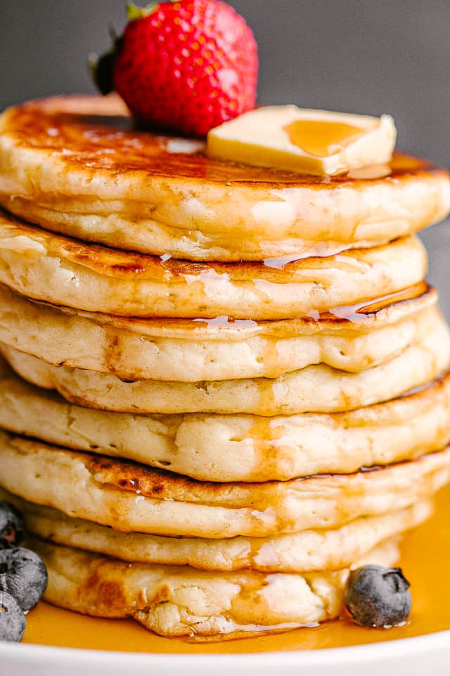 Fluffy pancakes stacked on top of each other, with a pat of butter and maple syrup for toppings. One strawberry is next to the pat of butter.