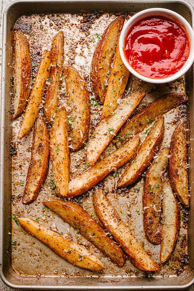 Crispy potato wedges on a baking sheet, with a cup of ketchup.