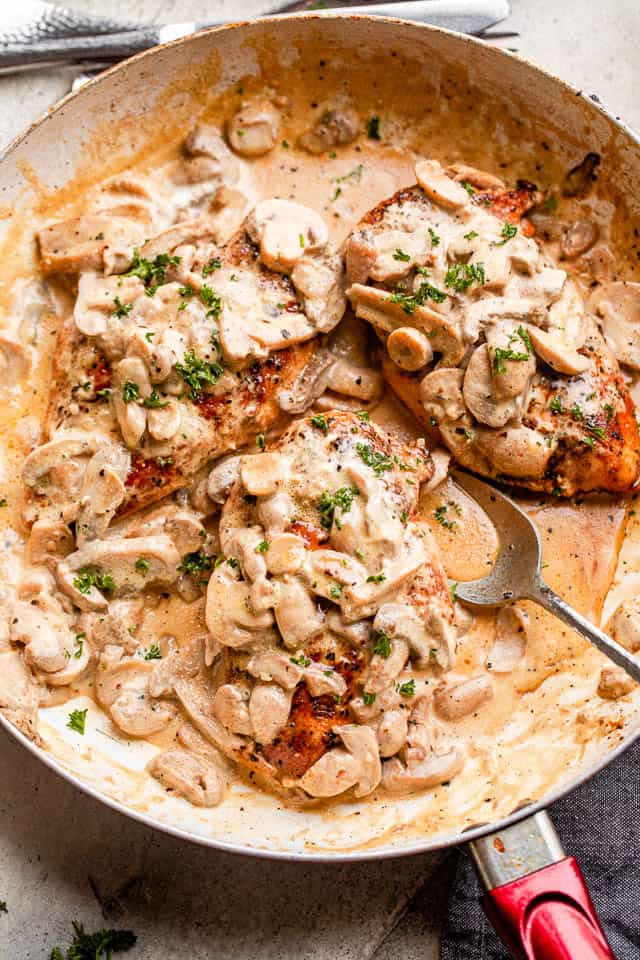 Golden-brown chicken breast in a skillet with creamy mushroom sauce, garnished with fresh herbs.