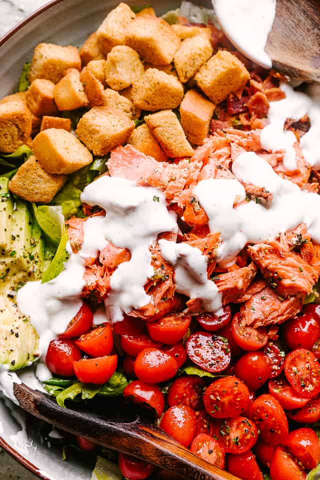 Croutons, flaked salmon, avocado, bacon, and other salad ingredients in a bowl, topped with ranch dressing.