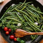 green beans, tomatoes, and garlic cloves in a black skillet with a wooden spoon inside