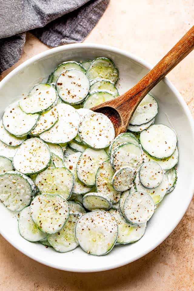 wooden spoon mixing cucumbers in a white salad bowl tossed with sour cream