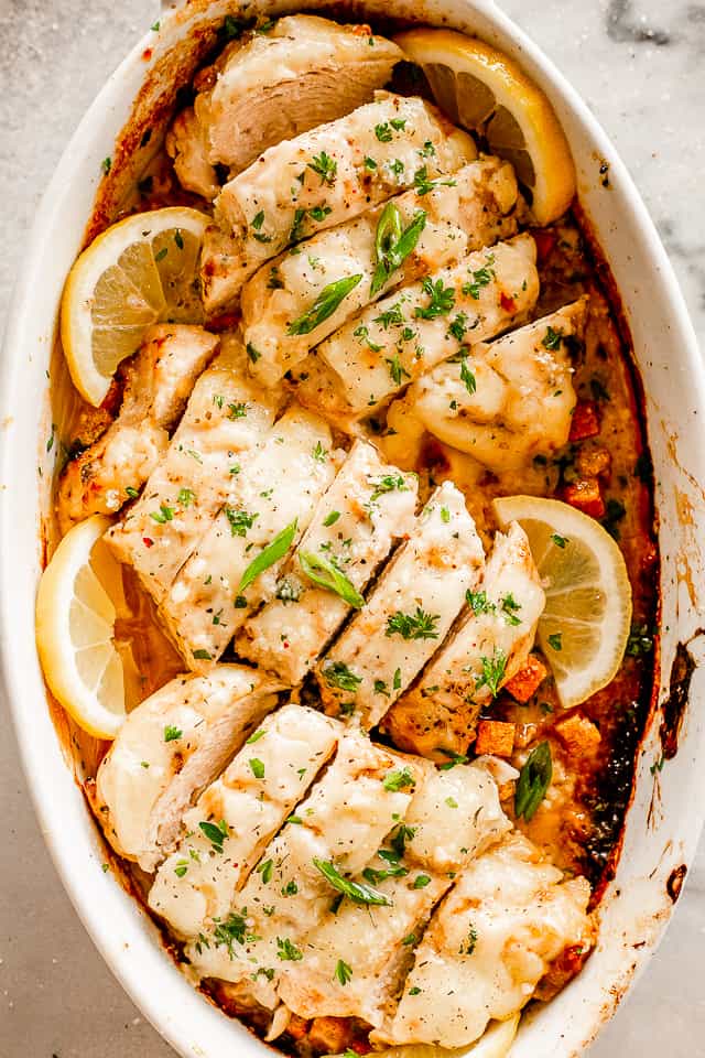 sliced caesar chicken breasts set in a white baking dish surrounded with lemon slices