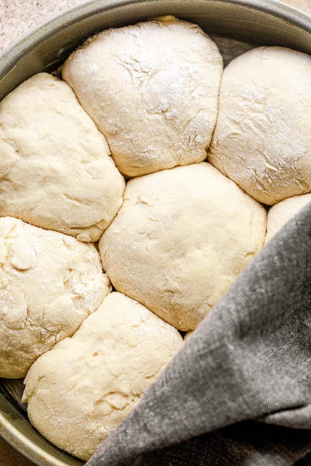 raw dinner rolls arranged in a cake pan and halfway covered with a grey kitchen towel