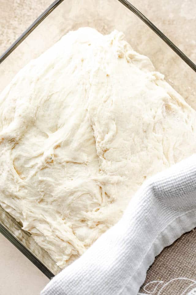 No Knead Dough in a glass container and halfway covered with a kitchen towel
