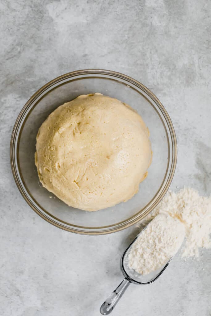 The Dinner Roll Dough in a Glass Bowl Beside a Scoop with Flour Inside It