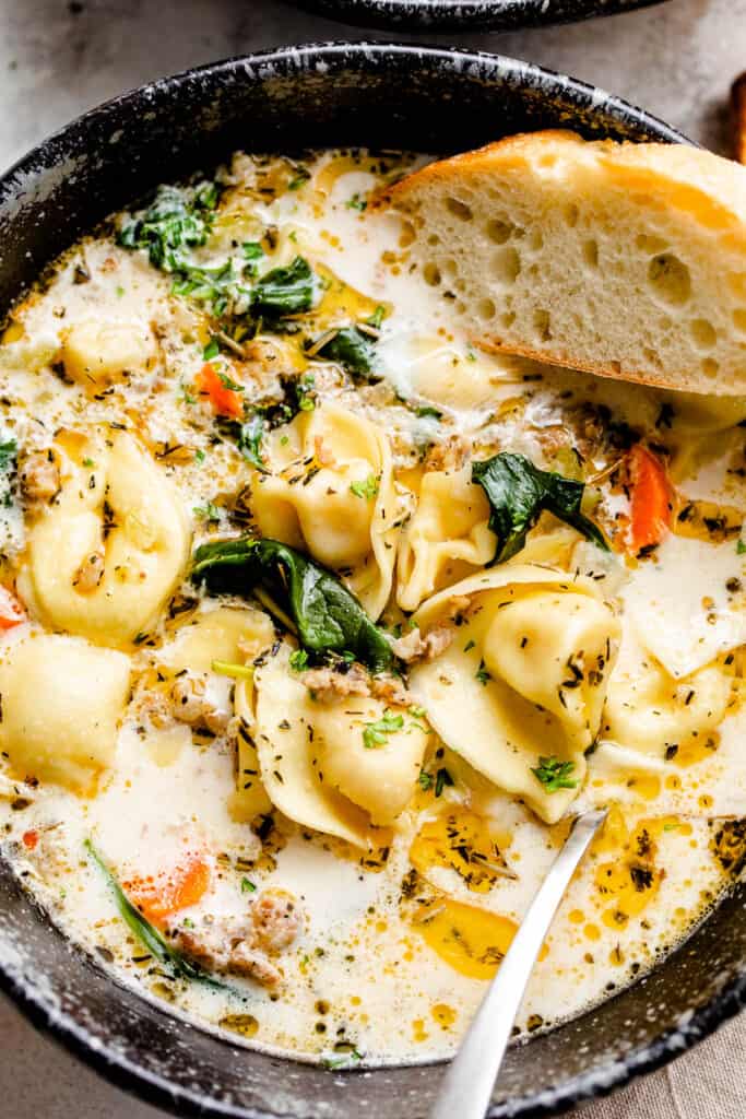 close up picture of creamy tortellini soup in a black soup bowl with slice of crusty bread