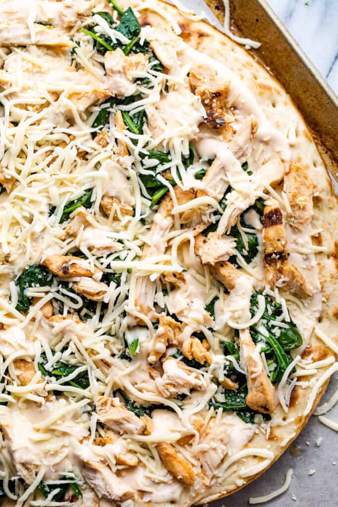 preparing pizza with alfredo sauce, shredded chicken, spinach, and cheese