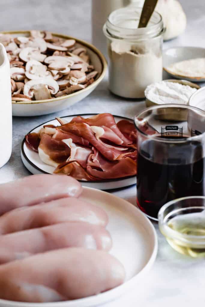 A plate of sliced white mushrooms, a plate of proscuitto, a cup of flour, and a cup of marsala wine.