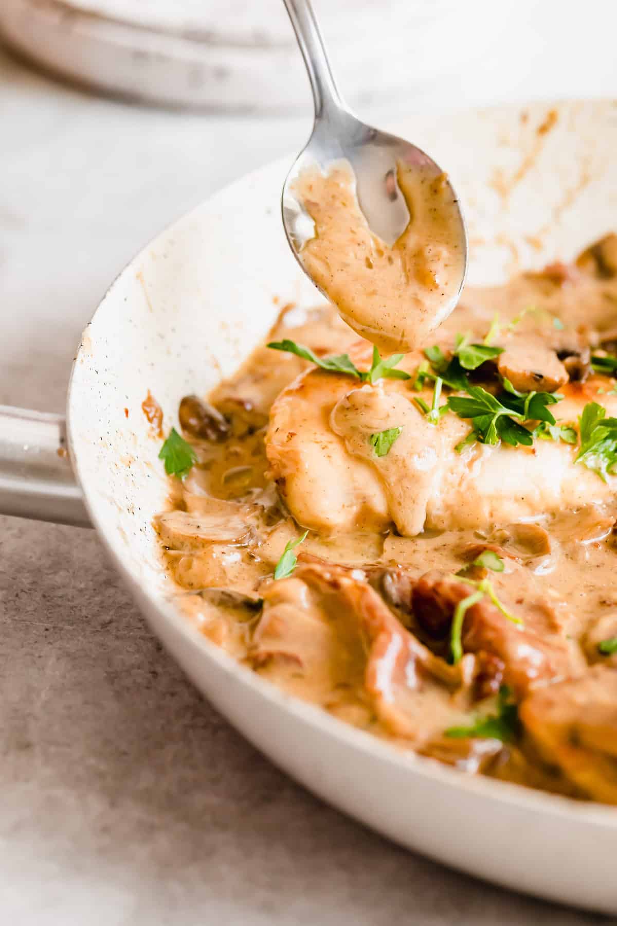 A spoon pouring creamy sauce over a piece of chicken with fresh herbs sprinkled on top.