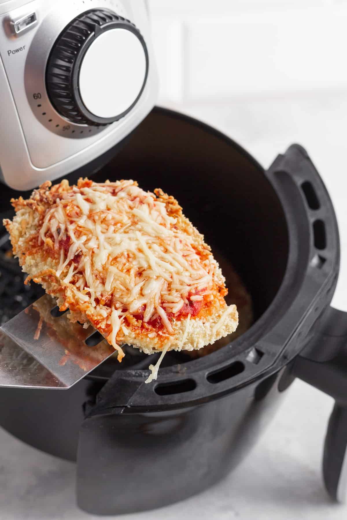 A Piece of Chicken Parm on a Metal Spatula Hovering Above an Air Fryer Basket