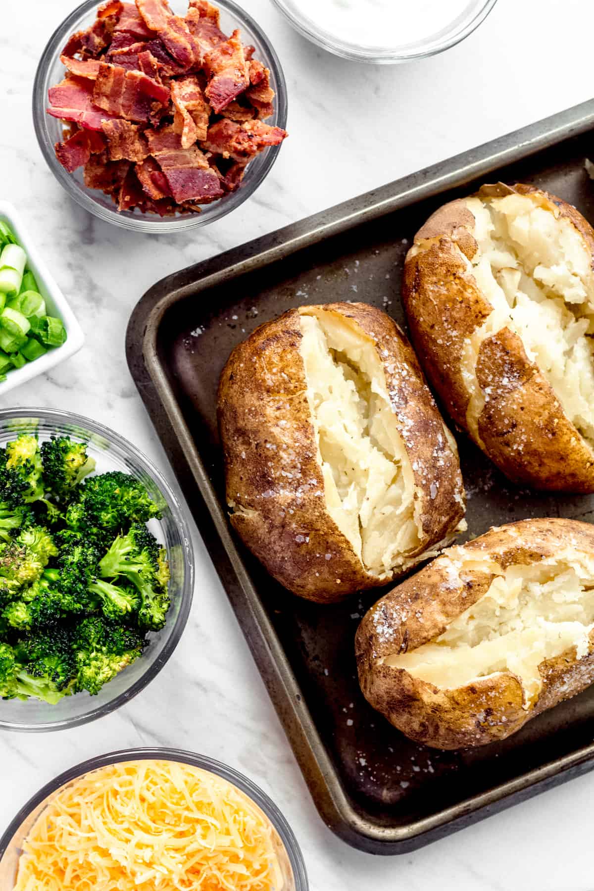 Three Instant Pot Baked Potatoes Beside Bowls of Cheese, Broccoli, Bacon, Chopped Green Onions and Sour Cream