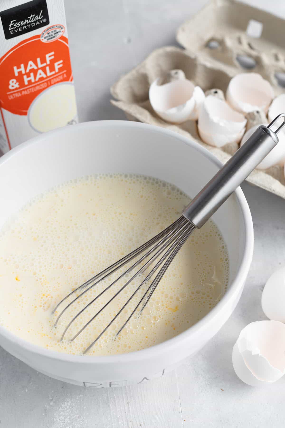 The Egg Mixture in a White Bowl with a Metal Whisk and Egg Shells in the Background