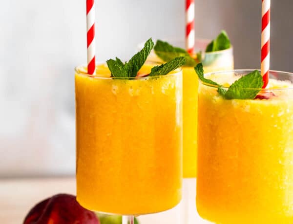 three glasses filled with peach daiquiris and garnished with mint, peach slices, and straws