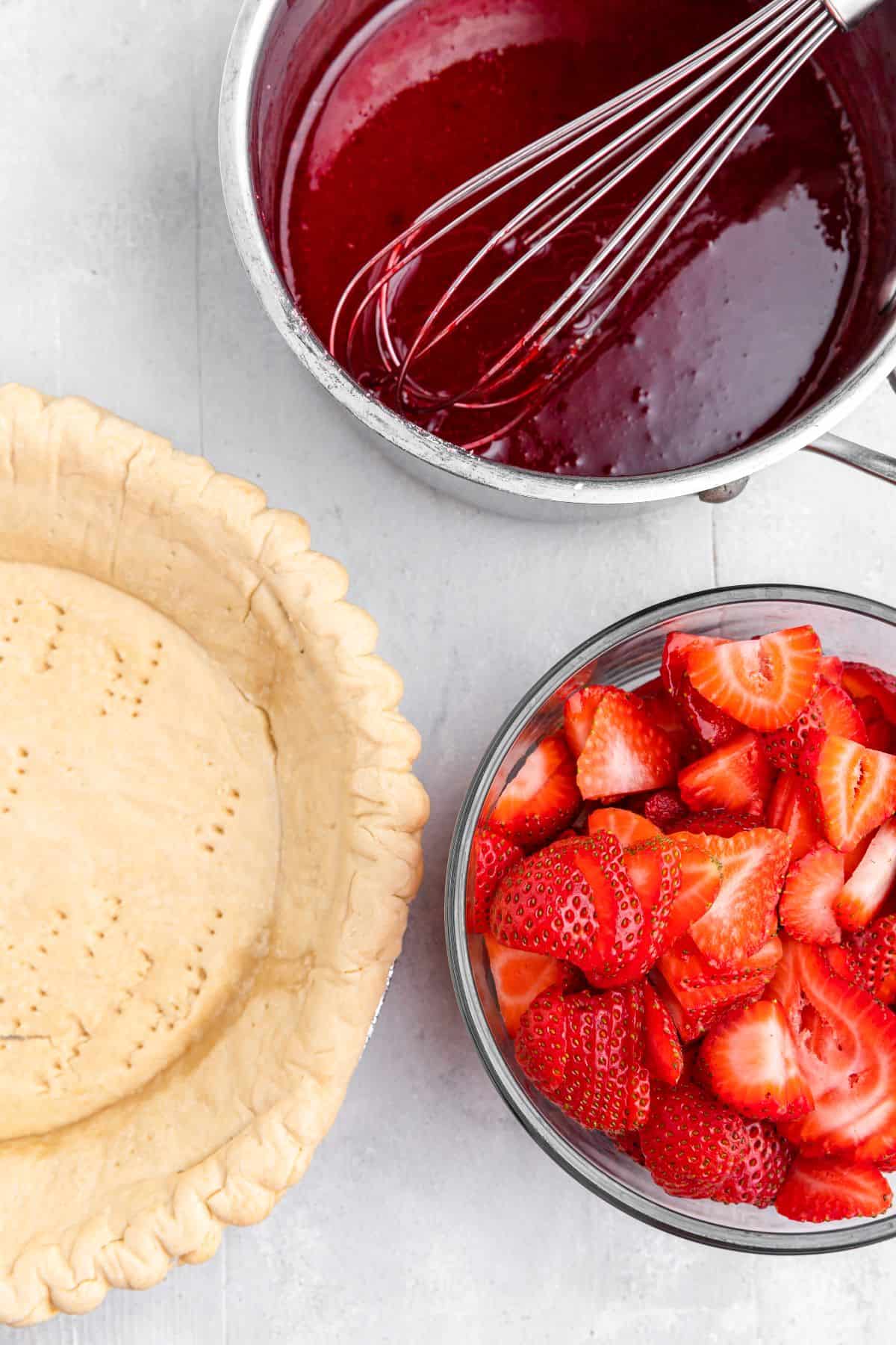 A pre-baked homemade pie crust, a bowl of fresh sliced strawberries, and a saucepan of warm strawberry gelatin on a work surface.