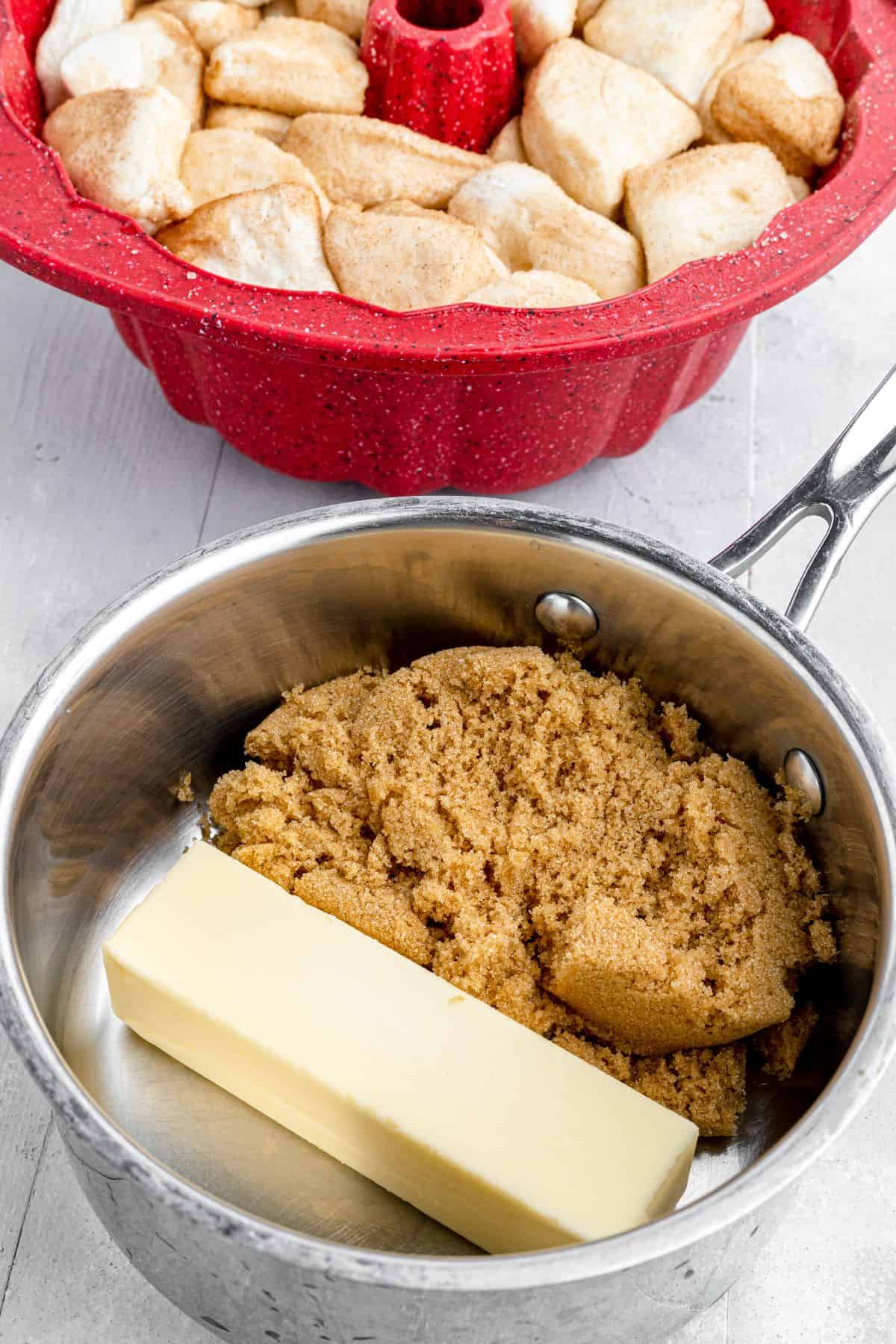 A Bundt pan filled with biscuit dough pieces sits next to a saucepan containing a stick of butter and some brown sugar.