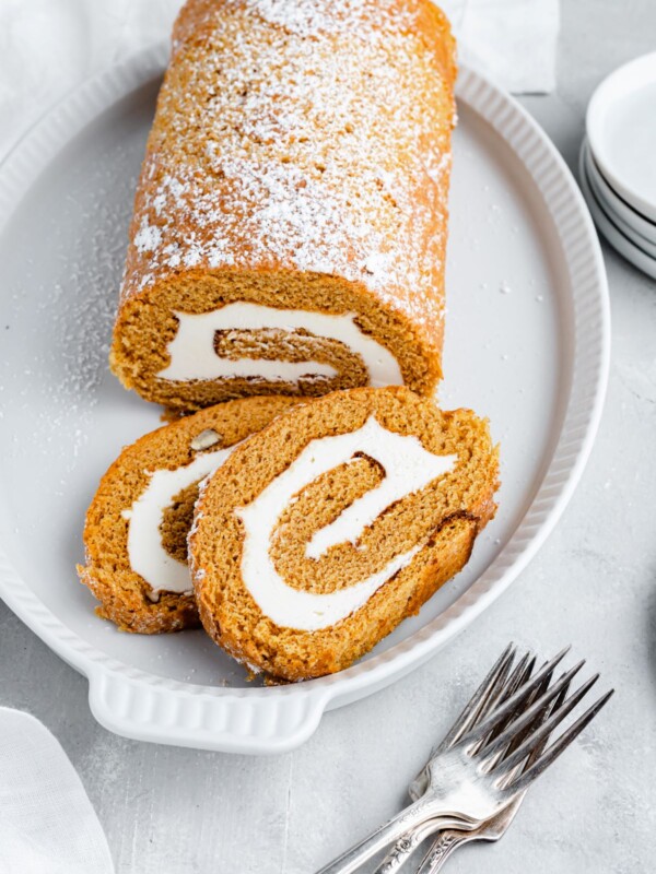 A pumpkin roll cake, dusted with powdered sugar, on a platter. Two slices have been cut and lie on the platter as well. Two forks are on the table nearby.