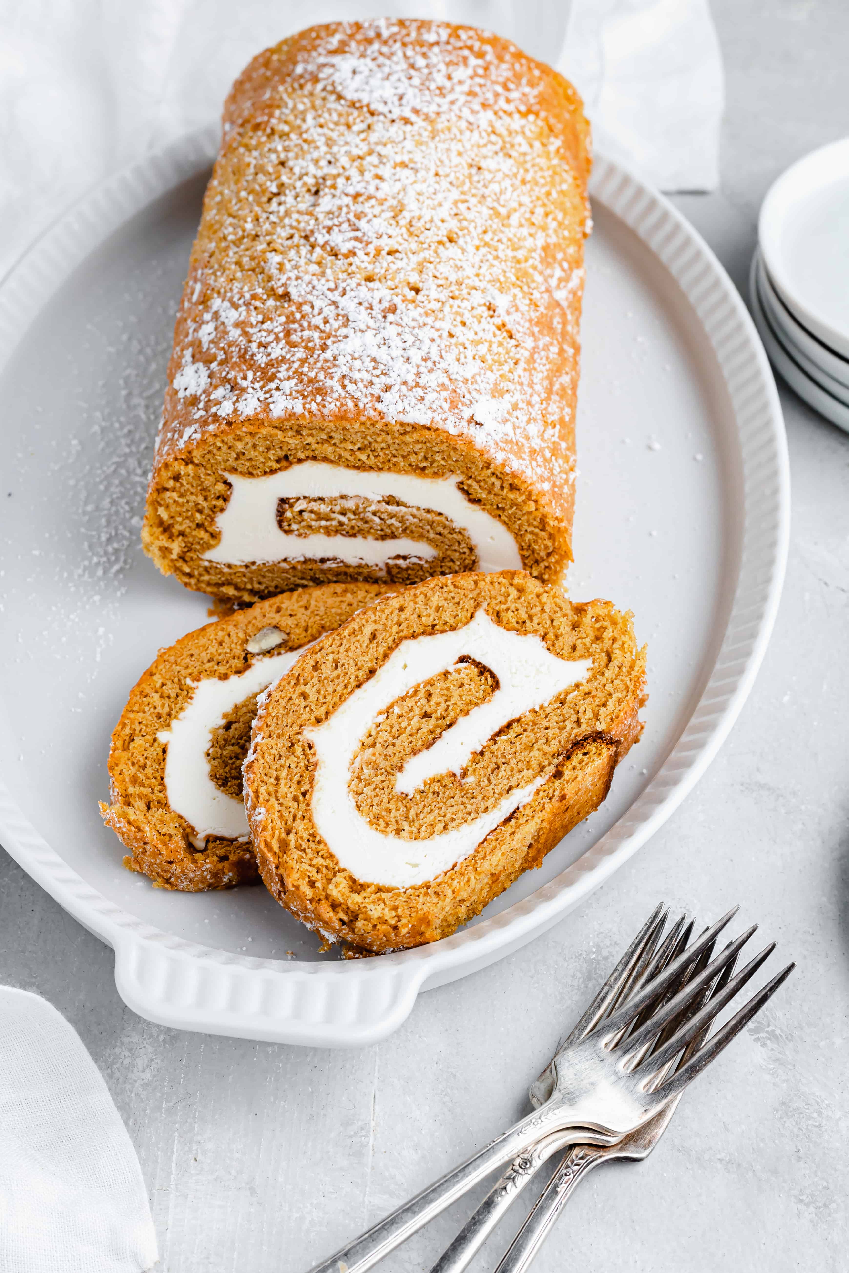 A pumpkin roll cake, dusted with powdered sugar, on a platter. Two slices have been cut and lie on the platter as well. Two forks are on the table nearby.