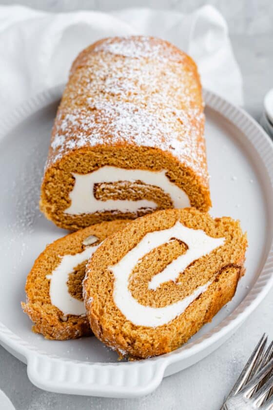 A shot of the pumpkin roll cake, dusted with powdered sugar, with two slices overlapping on a platter.