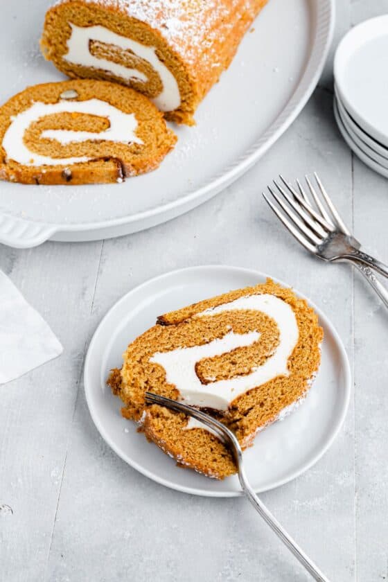 A slice of pumpkin roll cake with a fork cutting into it; in the background, the remaining cake is on a platter with another slice ready to serve.