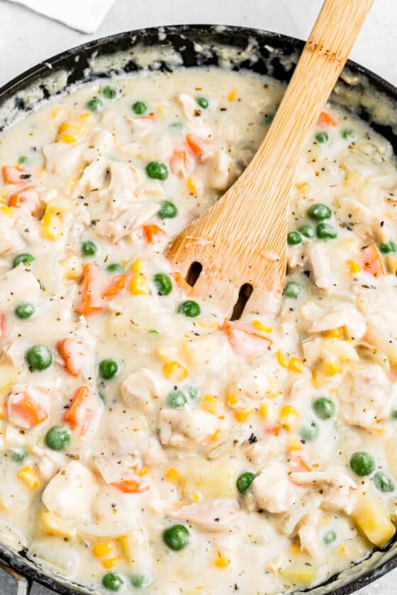 Creamy pot pie filling, featuring carrots, peas, corn, diced potatoes, and chicken, being stirred with a wooden spoon.