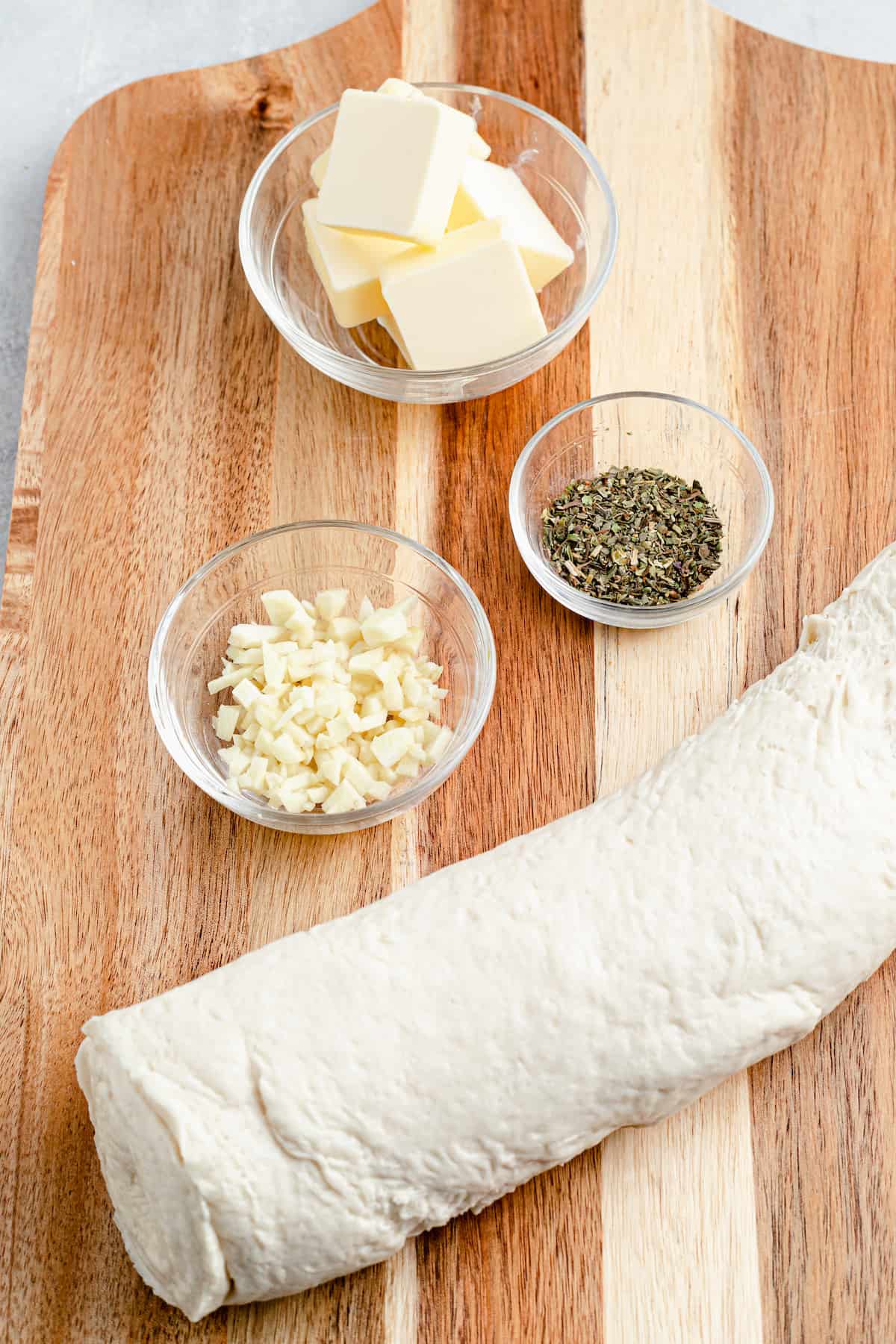 Clockwise from top: butter, Italian seasoning, a roll of pizza dough, a dish of minced garlic.