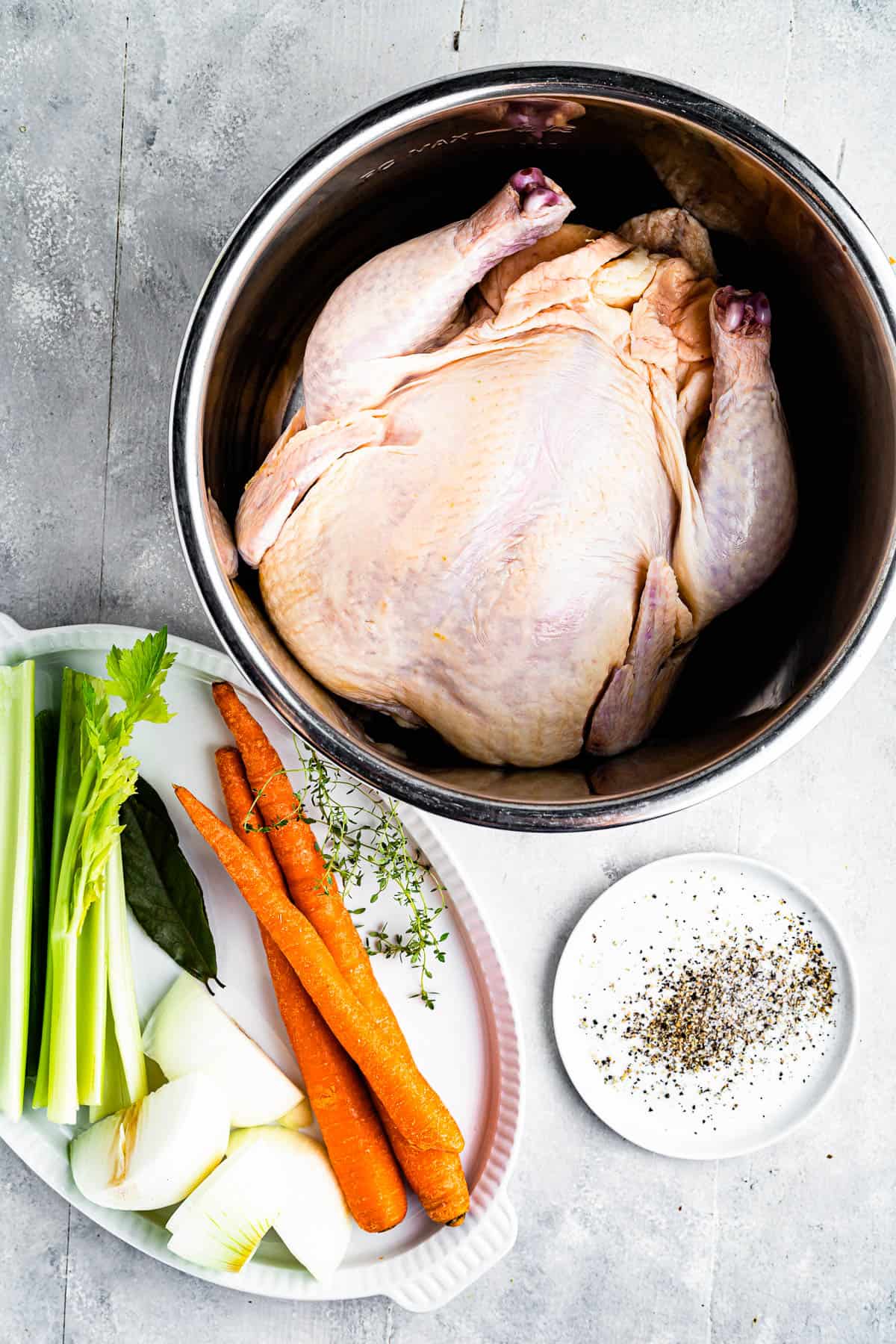 Clockwise from top: a whole chicken in a pot, seasonings, and a tray of raw vegetables including carrots, onions, and celery.