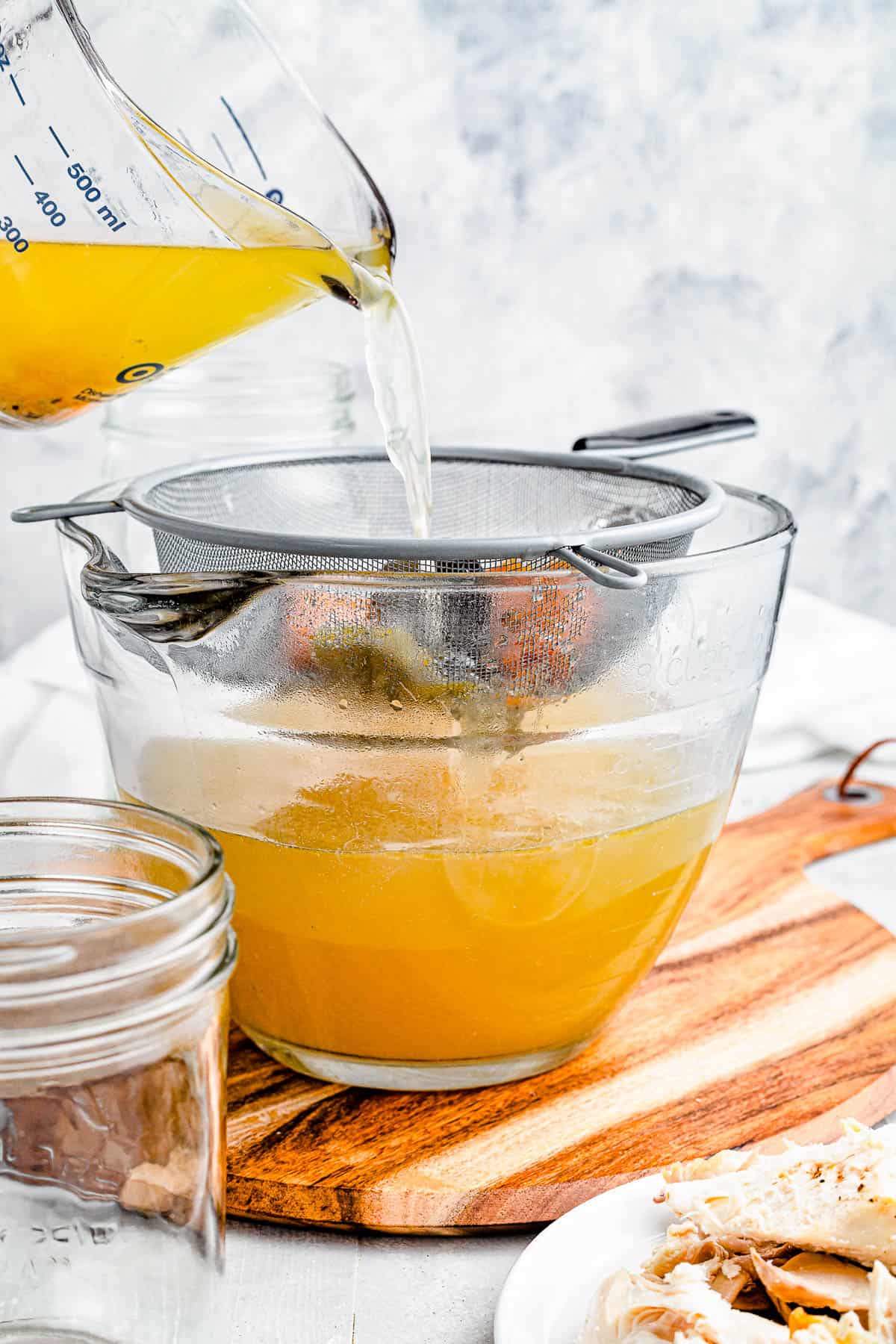 Chicken broth being poured from a large measuring cup into a bowl through a large sieve.