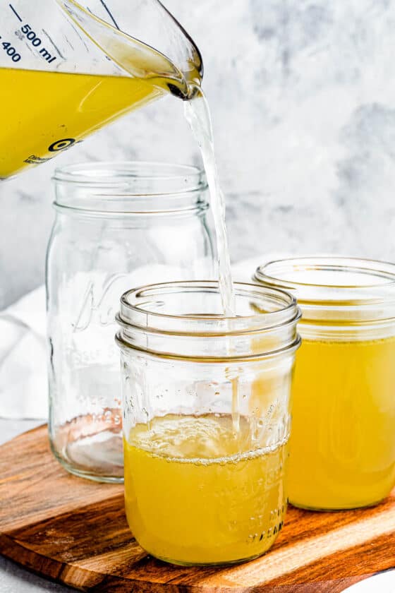 Chicken broth being poured from a large measuring cup into several jars.