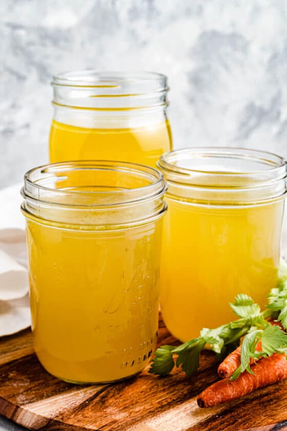 3 jars of homemade chicken broth on a cutting board with raw carrots nearby.