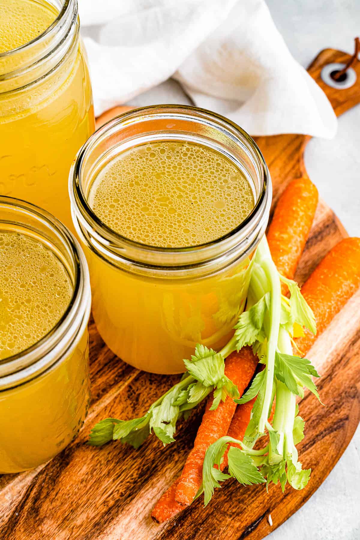 Three jars of chicken broth on a cutting board with raw carrots nearby.