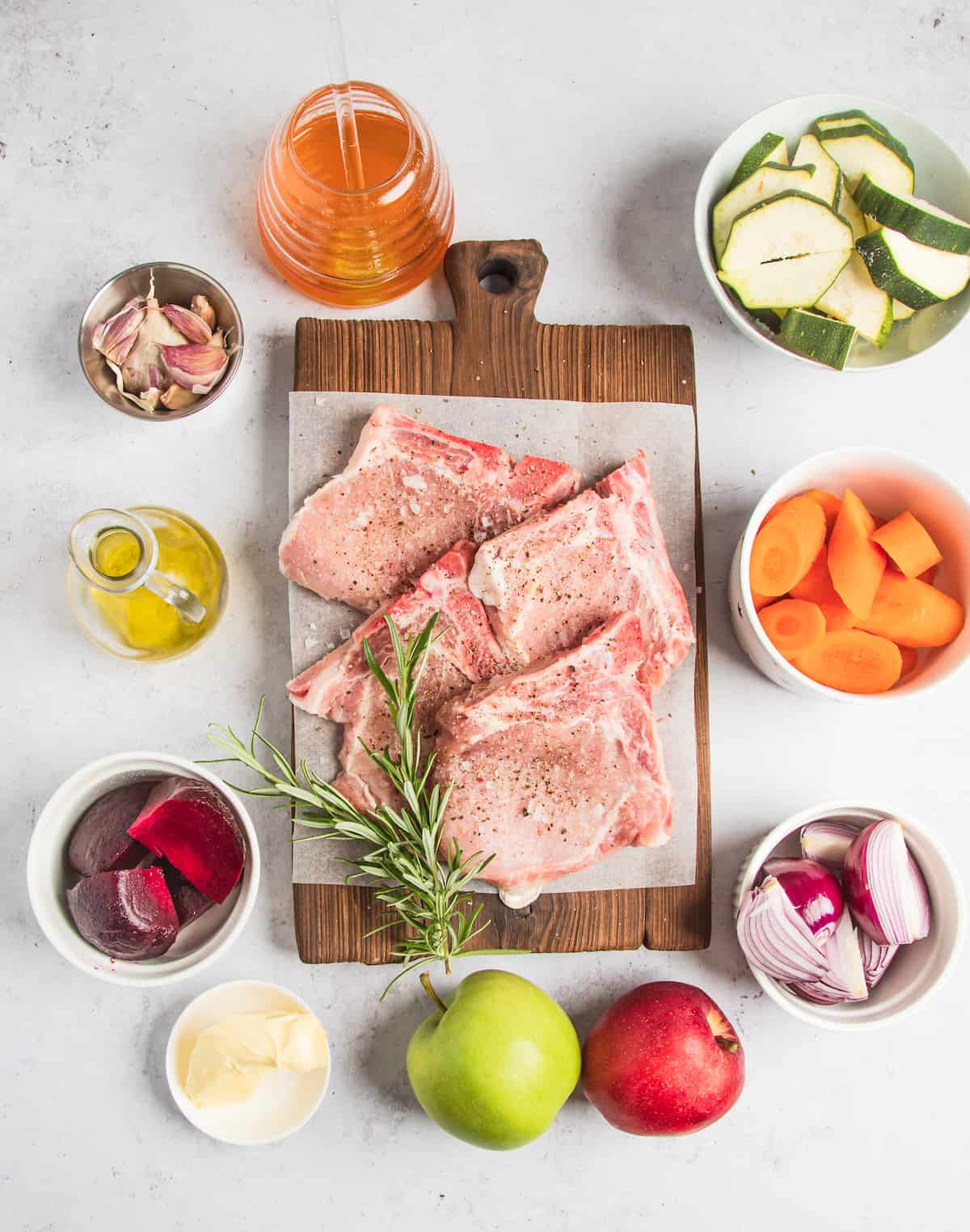 Clockwise from top: Sliced zucchini, chopped carrots, chopped onions, two whole apples, butter, beetroot, oil, garlic, and apple cider.  In the center: Four seasoned pork chops and a sprig of fresh rosemary.