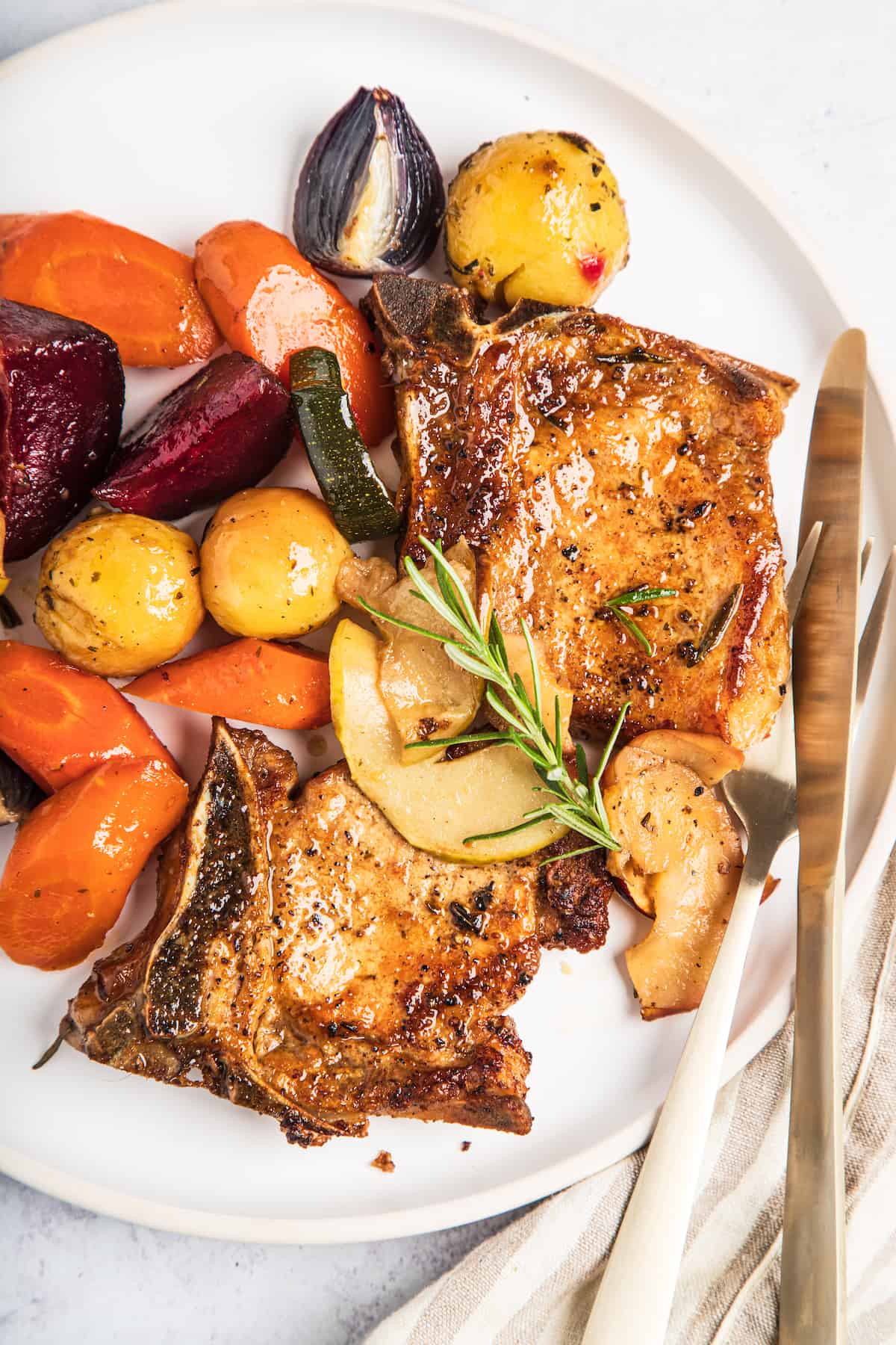 A plate of baked pork chops with apples and winter vegetables, garnished with a sprig of rosemary.