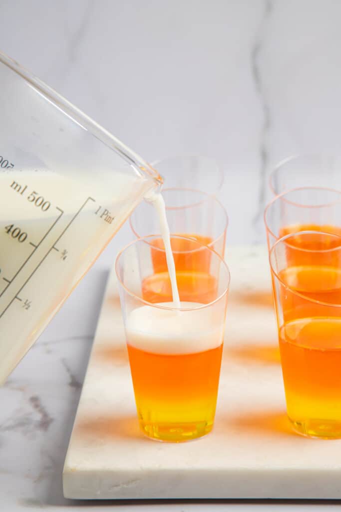 A creamy white layer being added to jello shot glasses on top of lemon and orange layers.