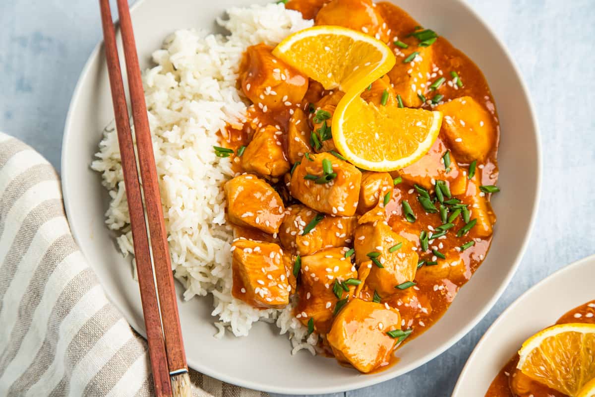a serving of orange chicken, garnished with orange wedges, on a plate with white rice and chopsticks.