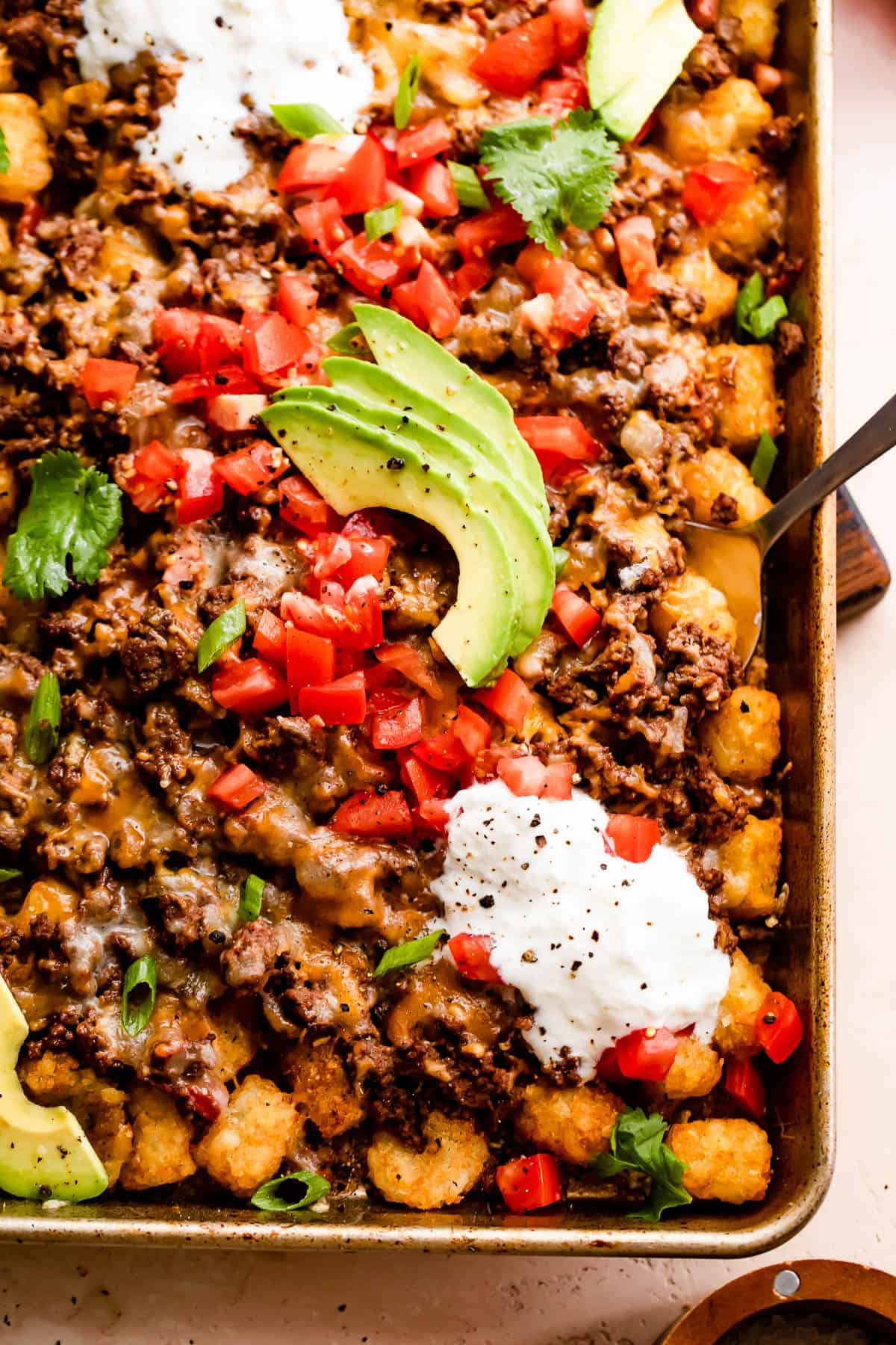 baking sheet with tater tots topped with ground beef, melted cheese, diced tomatoes, sour cream, and avocado slices
