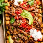 baking sheet with tater tots topped with ground beef, melted cheese, diced tomatoes, sour cream, and avocado slices