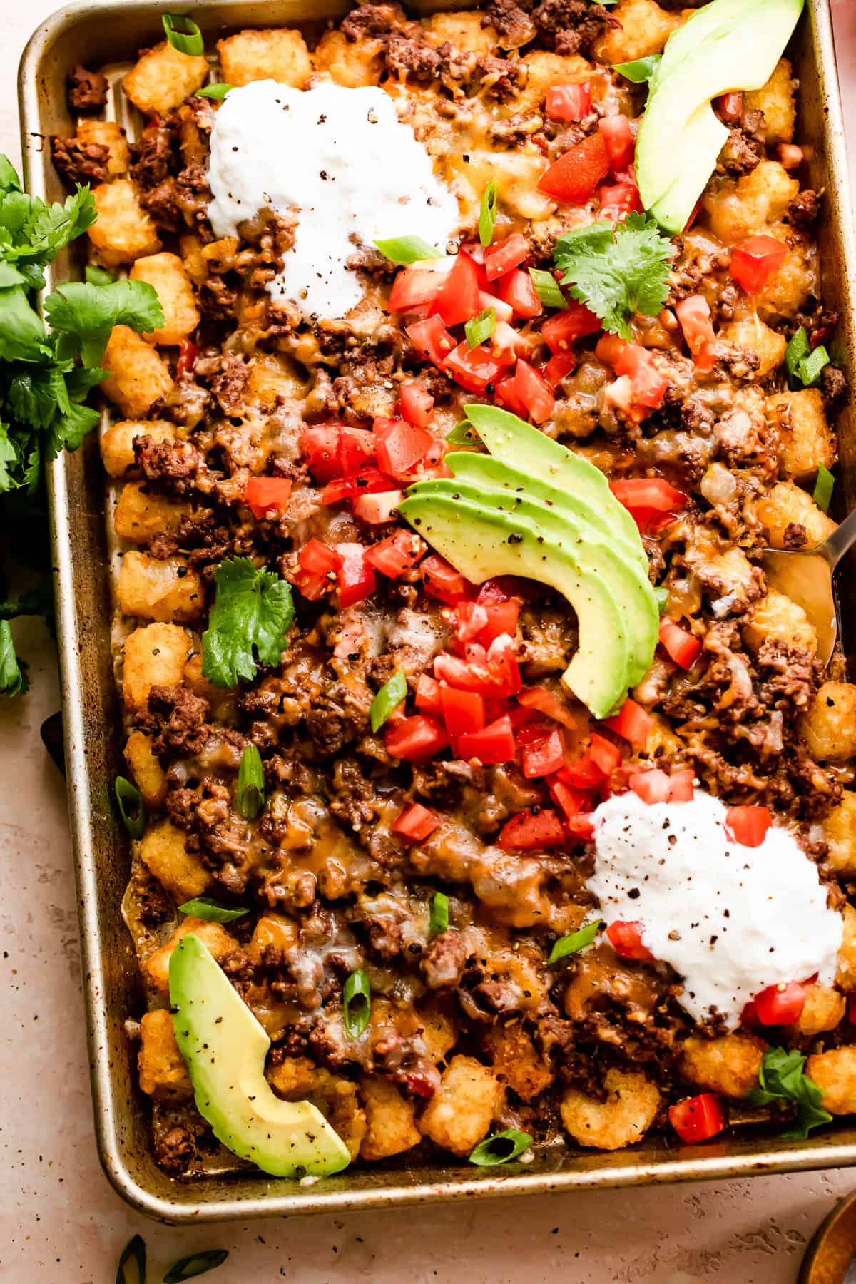 baking sheet with tater tots topped with ground beef, melted cheese, diced tomatoes, sour cream, and avocado slices