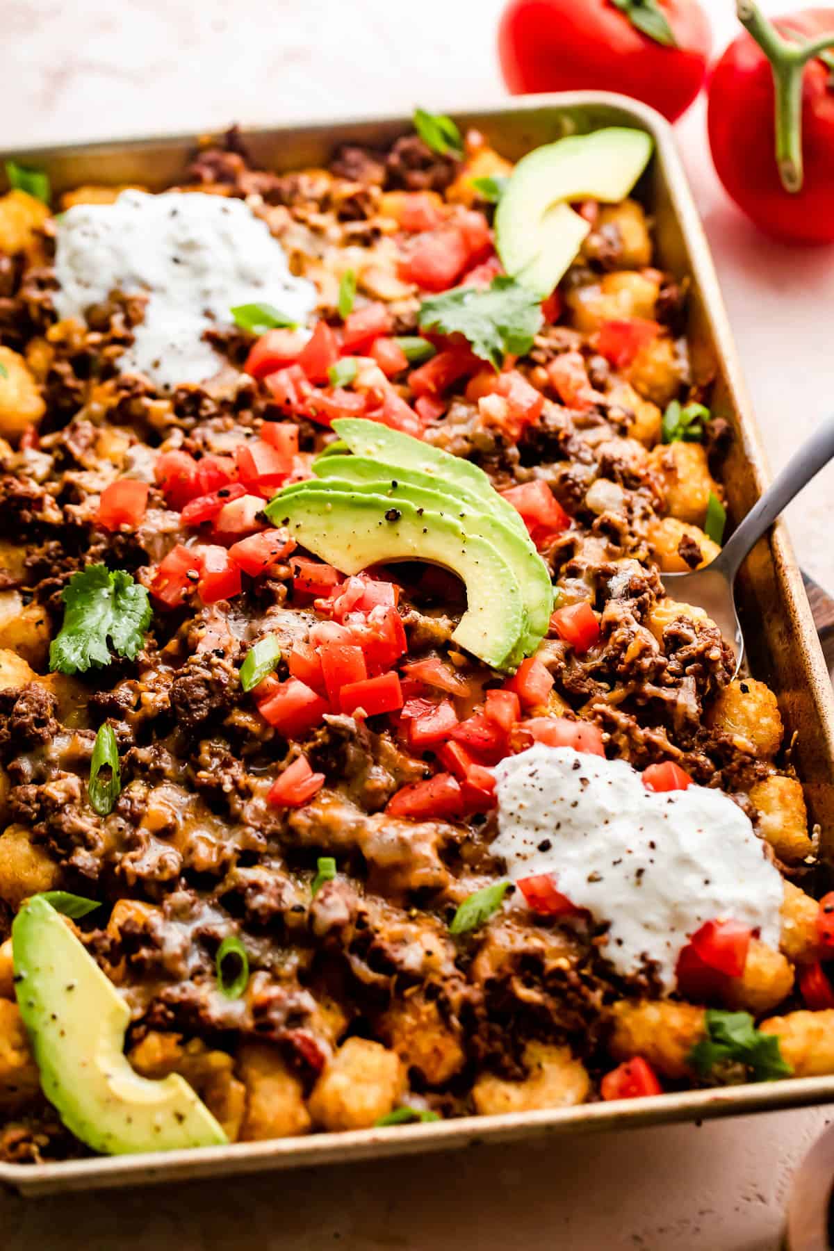 baking sheet with tater tots topped with ground beef, melted cheese, diced tomatoes, sour cream, and avocado slices