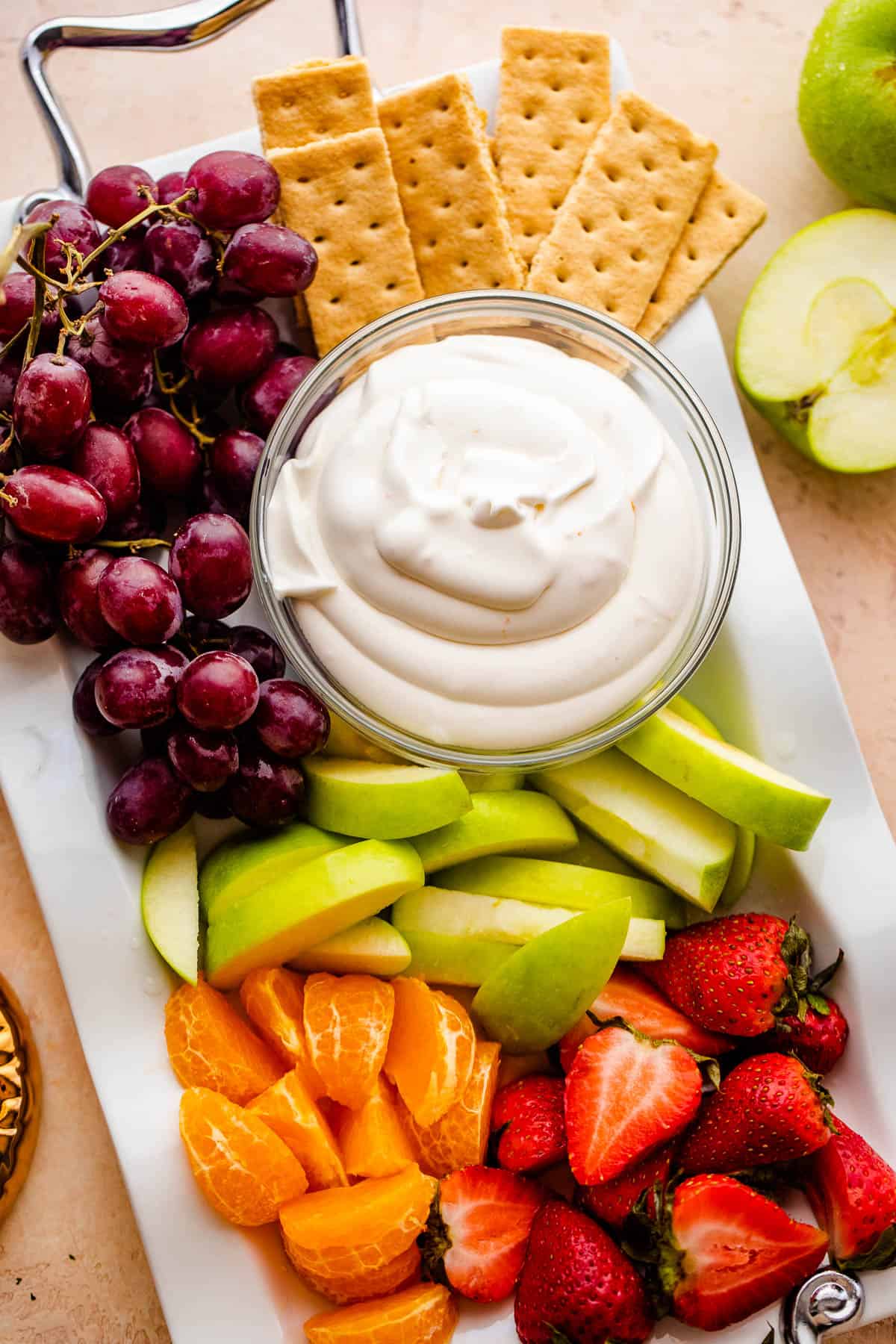 Bowl of creamy coconut fruit dip, green apple slices, tangerine slices, strawberries, grapes, and graham crackers arranged on a long serving plate. 