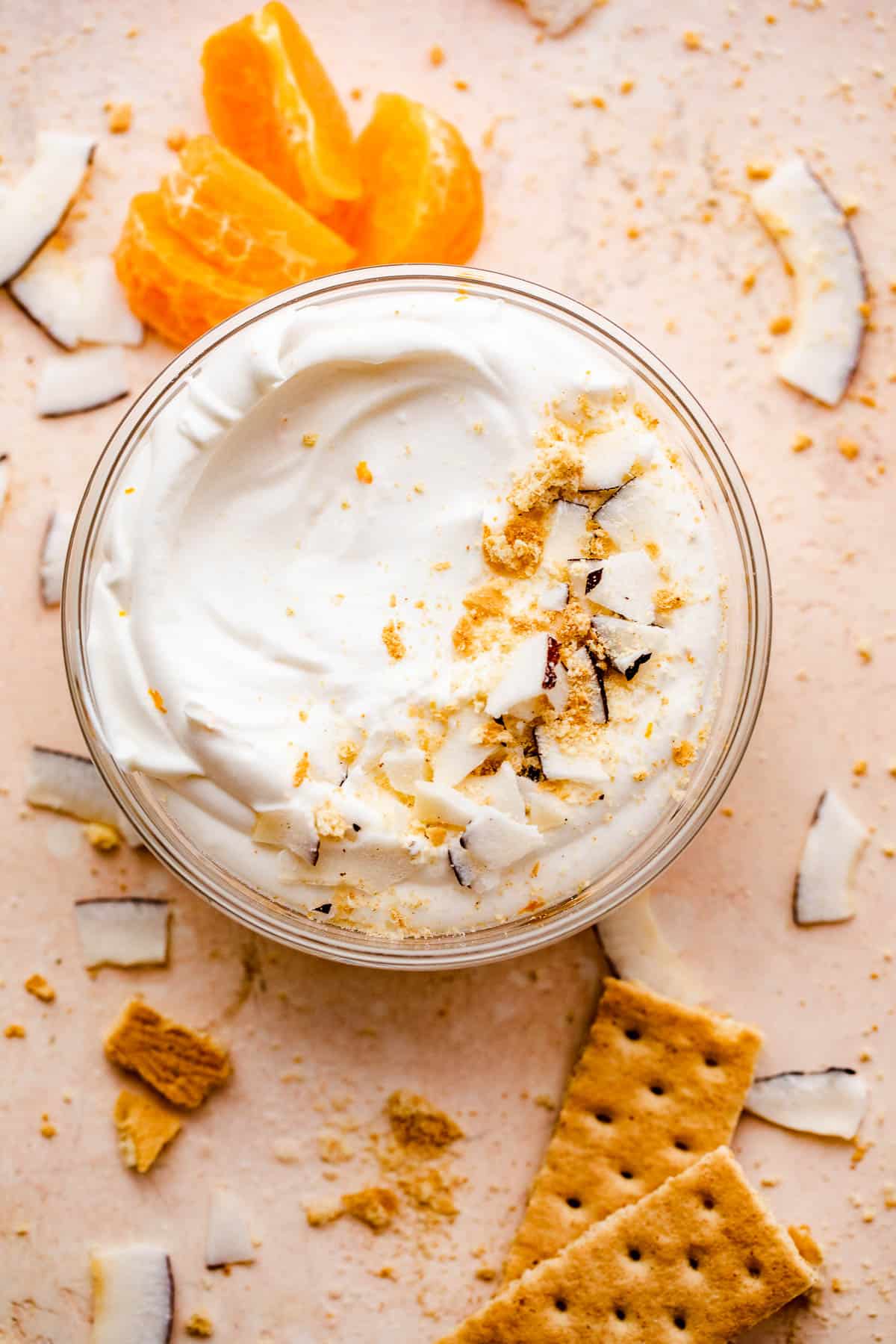 overhead shot of a creamy coconut dip served in a glass bowl with tangerine slices and coconut shavings placed around and on the sides of the bowl.