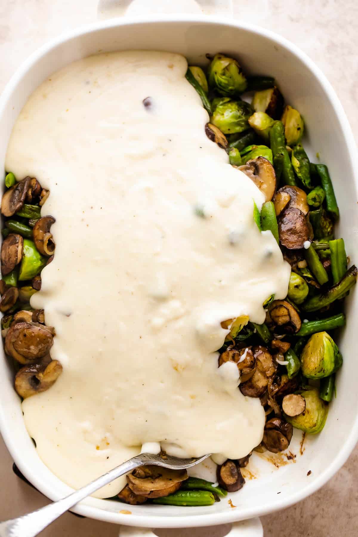 creamy sauce poured over brussel sprouts and green beans in a white baking dish
