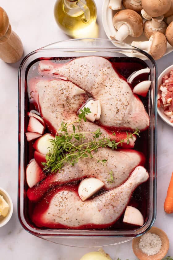A glass baking dish with chicken leg quarters, quartered onions, and fresh thyme, all marinating in red wine. The other coq au vin ingredients are arranged around the work surface.