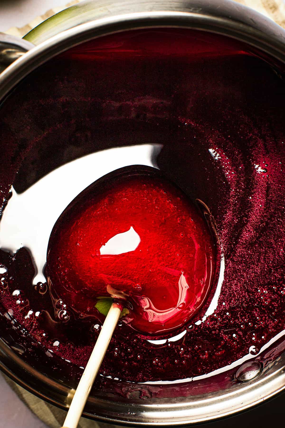 An apple coated in red candy is being lifted from a saucepan of the candy mixture.