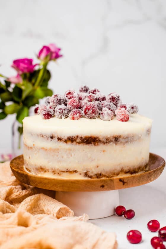 An assembled and iced cranberry carrot cake on a cake stand, topped with sugared cranberries.