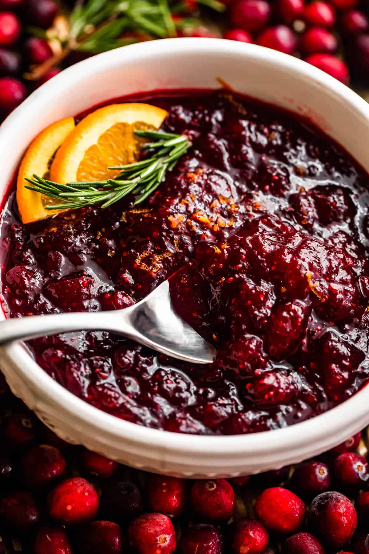 cranberry sauce inside of a white bowl surround by fresh cranberries and garnished with orange slices and rosemary sprig.