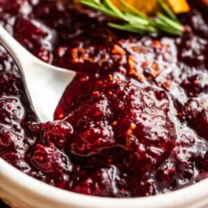 side shot of a spoon inside of a bowl filled with cranberry sauce and garnished with rosemary sprig plus orange slices