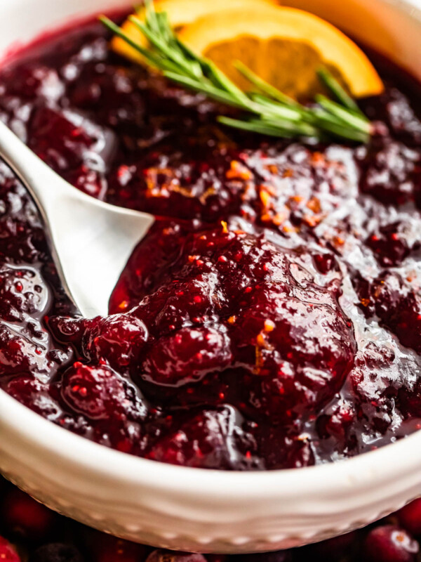 side shot of a spoon inside of a bowl filled with cranberry sauce and garnished with rosemary sprig plus orange slices